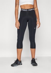 Leggings capri negros con una cinturilla ancha que presenta "PRO NIKE" en blanco. Textura suave con un pequeño logo de Nike en blanco en el muslo.