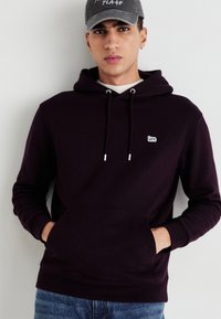 Felpa viola scuro con tasca a marsupio e coulisse, con un piccolo logo bianco. Indossata con jeans blu e un berretto grigio.