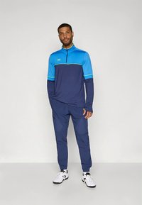 Modrý a námornícky športový outfit so zapínaním na zips a dlhými rukávmi, s ľahkými svetlomodrými akcentmi a ľahkou tkaninou. Biele športové topánky.