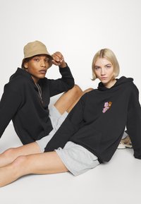 Black hoodies with front pockets; one features a colorful graphic. Grey shorts worn by models sitting against a white background.