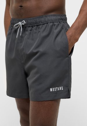 Man wearing black swim shorts with white "MUSTANG" text near hem, elastic waistband, and drawstring, standing with right hand in pocket.