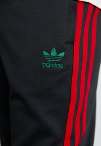 Svarta sportbyxor med röda sidoremsor. Har grönt broderat Adidas-logo, mjuk textur och åtsittande design.