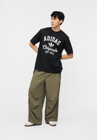 Νέος άνδρας που φοράει μαύρο μπλουζάκι Adidas Originals και φαρδιά χακί παντελόνια, στέκεται με τα χέρια πίσω από την πλάτη, κοιτώντας προς τα πλάγια.