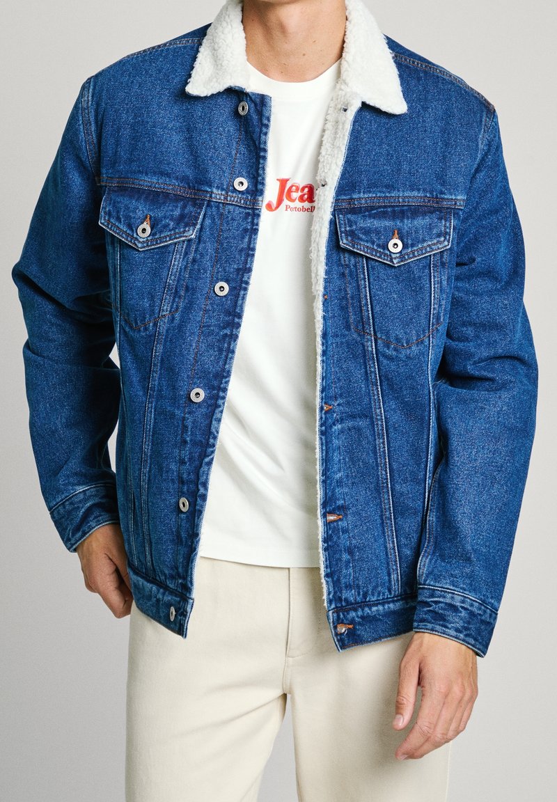 Jeansjacka med en ljusbrun sherpakrage, blåtvättad, med två bröstfickor och knappstängning. Bärs över en vit t-shirt.