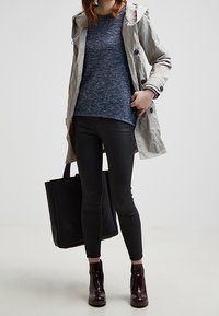 Un trench-coat gris clair porté sur un pull bleu texturé, associé à un jean slim noir et des bottines noires. Sac fourre-tout noir à la main.