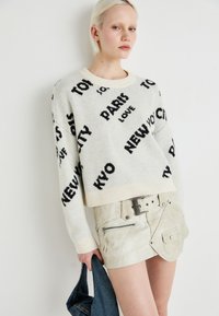 Hvit strikket genser med svarte tekstmønstre, cropped design. Kombinert med et beige skinnminiskjørt med belte og lommer.