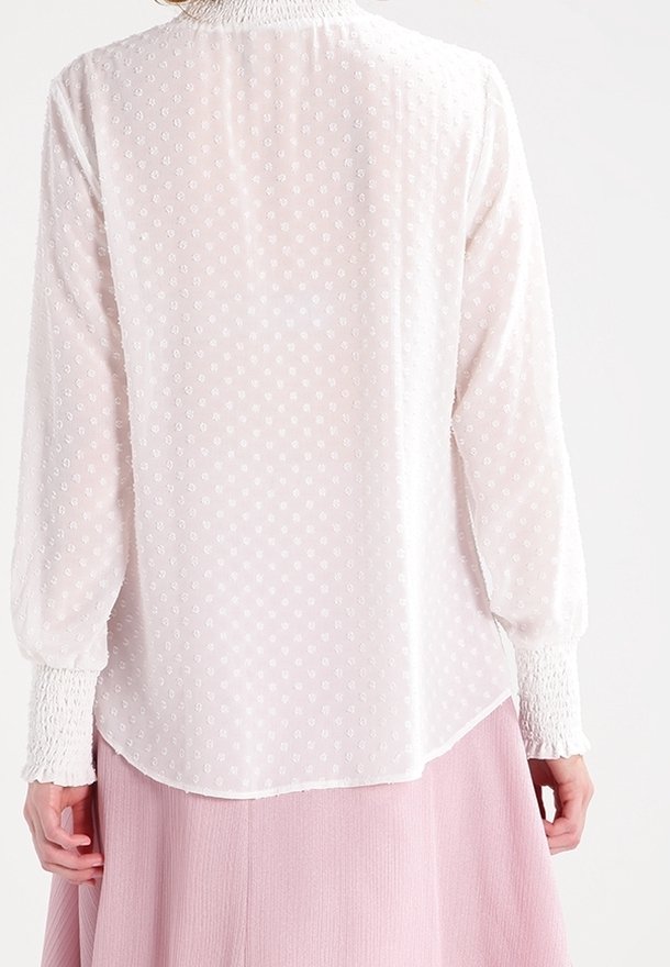 Personne portant un chemisier blanc transparent à manches longues avec des pois texturés et des poignets smockés, associée à une jupe plissée rose clair, vue de dos.