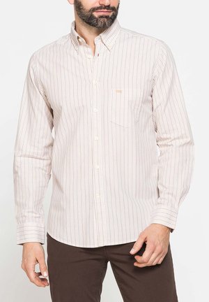 Uomo che indossa una camicia beige chiaro a maniche lunghe con sottili strisce verticali e pantaloni marrone scuro, in piedi davanti a uno sfondo semplice.