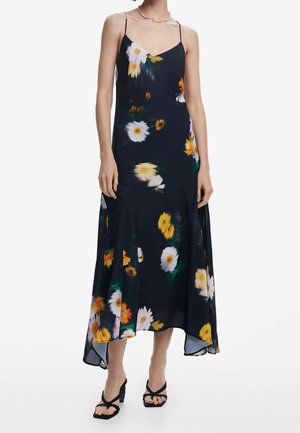Mujer con un vestido lencero negro con estampado floral, tirantes finos y dobladillo asimétrico, combinado con tacones abiertos negros.