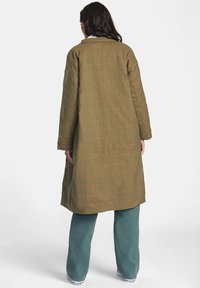 Abrigo largo de cuadros marrones con cuello, corte holgado y bolsillos laterales, combinado con pantalones verdes anchos y zapatillas. Se aprecia la textura del tejido.