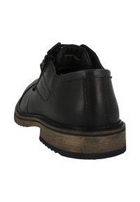 Chaussure en cuir noir avec une texture lisse, bout arrondi, et une semelle extérieure texturée de couleur marron clair contrastante. Présente une sangle décorative sur le côté.