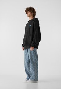 Sudadera negra de gran tamaño con bolsillo frontal y logo azul. Vaqueros azules de corte holgado con un diseño geométrico y zapatillas blancas.