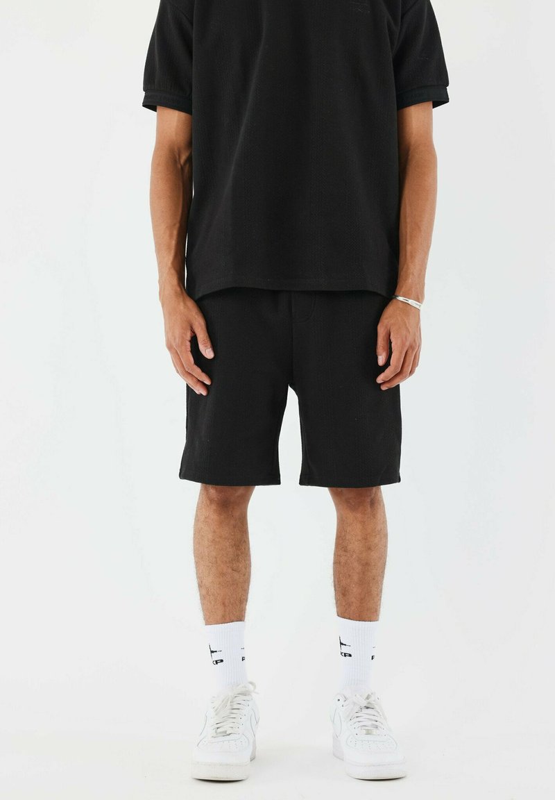 Camiseta de manga corta negra y pantalones cortos hasta la rodilla de tejido texturizado, combinados con calcetines blancos y zapatillas deportivas. Diseño simple, color monocromático.