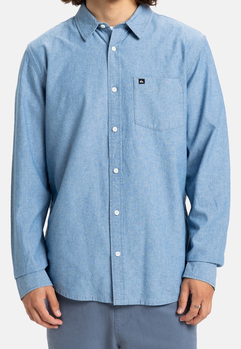 Homme portant une chemise bleu clair à manches longues avec une poche poitrine et une petite étiquette noire avec logo sur la poche.