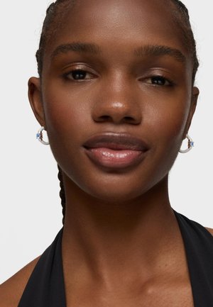 Primo piano di una donna con i capelli intrecciati che indossa orecchini a cerchio in argento con gemme blu e un top halter nero su uno sfondo neutro.