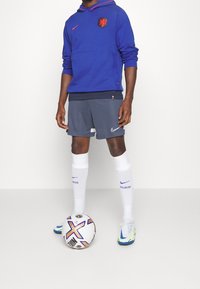 Felpa con cappuccio blu e dettagli rossi, pantaloni corti grigi, calze bianche e scarpini da calcio multicolori. Un pallone da calcio a terra.