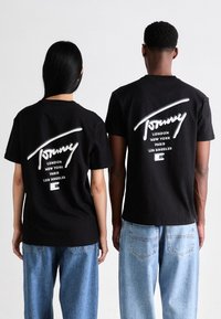 Kaksi ihmistä seisoo selin päin, päällä mustat Tommy Jeans -t-paidat, joissa on kaupunkien nimet Lontoo, New York, Pariisi, Los Angeles, yhdistettyinä vaaleansiniin farkkuihin.