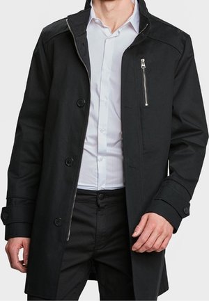 Homme portant un manteau noir boutonné avec une poche zippée, par-dessus une chemise blanche et un pantalon noir, visible du cou jusqu'à mi-cuisse.