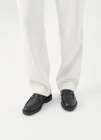 Schwarze Leder Slipper mit glatter Oberfläche, abgerundeter Spitze und Akzentnähte. Kombiniert mit weit geschnittenen weißen Hosen.