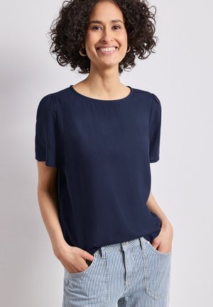 Usměvavá žena s kudrnatými vlasy, nosící zlaté kruhové náušnice, tmavě modrou blůzu s krátkými rukávy a modro-bíle pruhované džíny s rukama v kapsách.