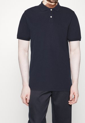 Muž v černém polo tričku s krátkými rukávy a dvěma bílými knoflíky, oblečený do tmavých kalhot, stojí před jednobarevným pozadím.