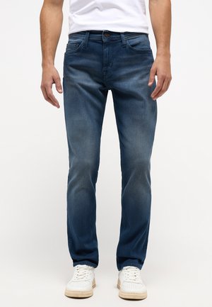Jeans de mezclilla azul oscuro con corte recto, desgastes visibles, diseño de cinco bolsillos y cierre de botón. Combinados con zapatillas deportivas blancas.