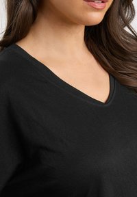 Donna con lunghi capelli castani che indossa una maglia nera a scollo a V, mostrando il collo, la parte inferiore del viso e parte della spalla.