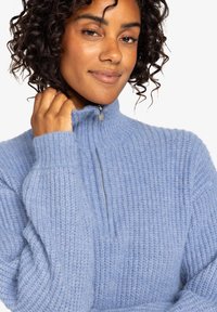 Světle modrý, žebrovaný pletený svetr s vysokým límcem, polovičním zipem a dlouhými rukávy. Měkká textura s uvolněným střihem.