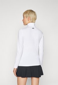 Λευκή αθλητική μπλούζα με μακριά μανίκια και ψηλό γιακά, με ένα μικρό μαύρο λογότυπο στην πλάτη, συνδυασμένη με μια πλισέ μαύρη φούστα.