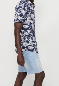 Homme portant une chemise bleu marine à manches courtes avec un imprimé tropical blanc et un short en jean délavé bleu clair, se tenant devant un fond pâle.