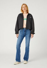 Svart croppad hoodie med dragkedja, snörmidja och elastiska ärmar, bärs över en rosa topp med gröna bokstäver, ihop med blå utsvängda jeans.