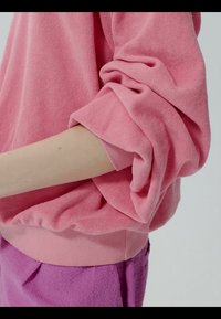Bras portant un pull rose à manches larges retroussées et un pantalon violet texturé, sur un fond uni.