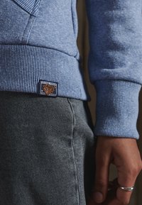 Sudadera azul con puños acanalados, que presenta una tela texturizada. Incluye una etiqueta cuadrada con un gráfico de un tigre y texto cerca del dobladillo. Jeans grises.