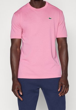 Dunkelhäutige Person trägt ein rosa T-Shirt mit kurzen Ärmeln, auf dem ein kleines grünes Krokodil-Logo zu sehen ist, und dunkelblaue Hose, steht vor weißem Hintergrund.