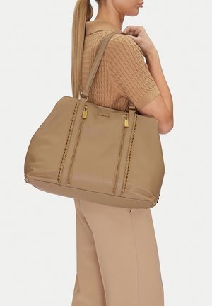 Donna con coda di cavallo lunga che indossa un top a maglia maniche corte color sabbia e pantaloni, che porta una grande borsa tote in pelle beige con borchie e cerniere dorate.