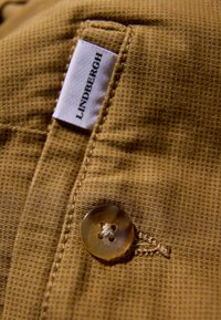 Tissu marron texturé avec un bouton visible et des coutures. Une étiquette portant l'inscription « LINDENBERGH » est attachée près du bouton.