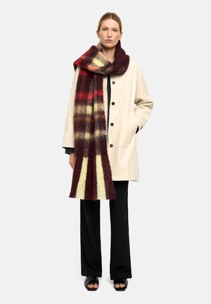 Beżowy płaszcz z ciemnymi guzikami, stylizowany na czarny top i luźne czarne spodnie. Czerwona i żółta kraciasta szalik luźno opasana wokół szyi.