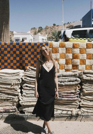 Czarna sukienka z dekoltem w kształcie litery V, stojąca przed ułożonymi tekstyliami. W tle kolorowy wzór w kratę na tkaninie.