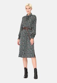 Robe à motif floral avec une base verte, des manches longues, un col à boutons et une ceinture marron. Longueur genou avec fentes latérales. Bottines noires.