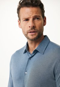 Ljusblå skjorta med knäppning i strukturerad tyg, klassisk krage och kontrasterande knappar, med en subtil gradienteffekt.