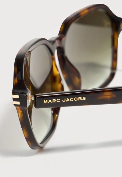 Γυαλιά ηλίου με καφέ φίδερα και οβάλ φακούς, με χρυσό λογότυπο "MARC JACOBS" στο πλαϊνό μπράτσο, σε λευκό φόντο.