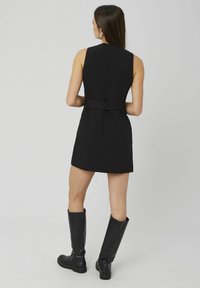 Robe noire sans manches au design ajusté, taille cintrée, avec un détail de bouton à l'arrière, accompagnée de bottes noires montantes.