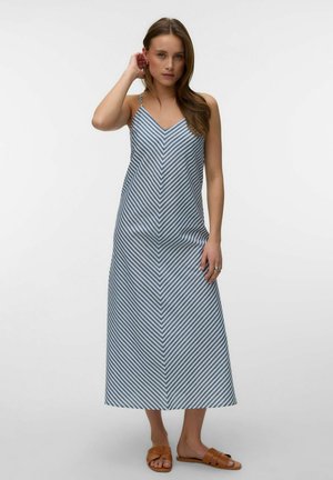 Kvinde iført en ærmeløs, V-hals, blå og hvide stribet maxi-kjole med brune flade sandaler, stående mod en ensfarvet lys baggrund.