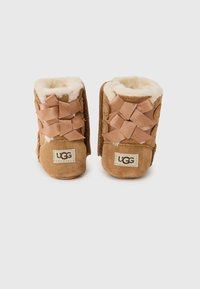Bruna mockaskor för småbarn med shearlingfoder, med korslagda band på framsidan och en liten broderad UGG-etikett på sidan.