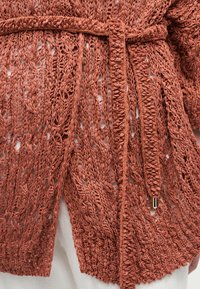 Λεπτομερής πλεκτή ζακέτα σε σκουριά απόχρωση με περίπλοκο ανοιχτό πλεκτό σχέδιο και ζώνη δεμένη στη μέση πάνω από λευκά ρούχα.