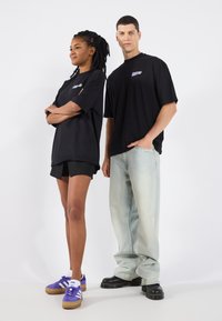 Sort oversized t-shirts med et lille multicolor logo, parret med lyseblå jeans og mørke sko. Modellen har shorts og lilla sneakers på.