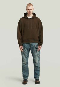 Donkerbruine hoodie met een kangoeroezak, gecombineerd met lichtblauwe cargobroek met meerdere zakken en een relaxed fit. Zwarte laarzen maken de look af.