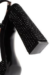 Scarpa con tacco alto nera con un tacco a blocco decorato in nero e texture. La tomaia presenta una finitura lucida nera e un dettaglio con cinturino alla caviglia.