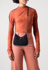 Bolso negro acolchado con letras en relieve, correa de cadena plateada, combinado con una blusa de manga larga color óxido y pantalones melocotón. Diseño simple.
