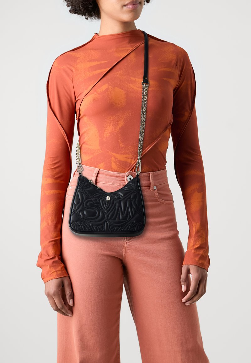 Bolso negro acolchado con letras en relieve, correa de cadena plateada, combinado con una blusa de manga larga color óxido y pantalones melocotón. Diseño simple.